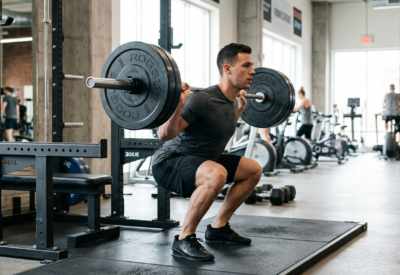 Man voert barbell squat uit met correcte techniek in een trainingshal met natuurlijk licht.